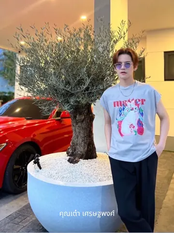 Tao Sattaphong Phiangphor, wearing glasses, stands beside an olive tree in a white planter.